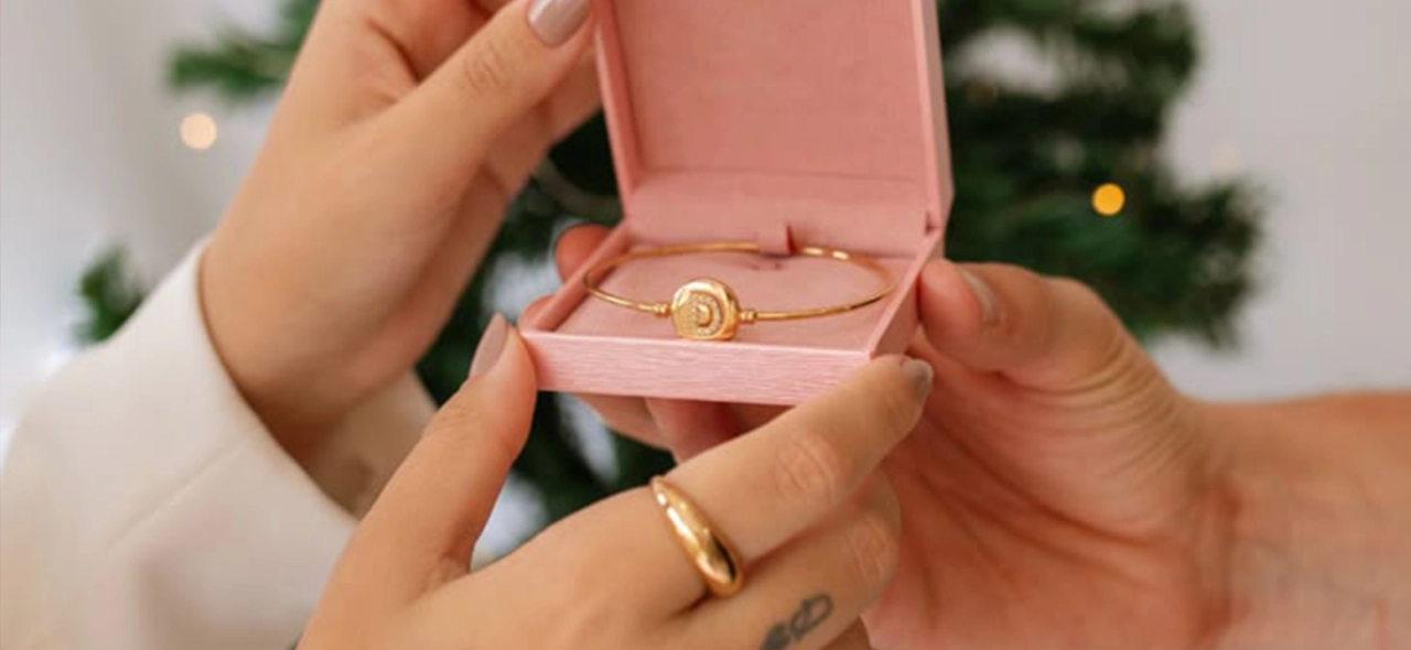 Mãos entregando uma pulseira dourada em caixa rosa diante de uma árvore de Natal